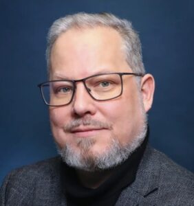 Älterer Mann mit Brille und grauem Vollbart, lächelnd, mit schwarzem Rollkragen und grauem Sakko vor blauem Hintergrund.