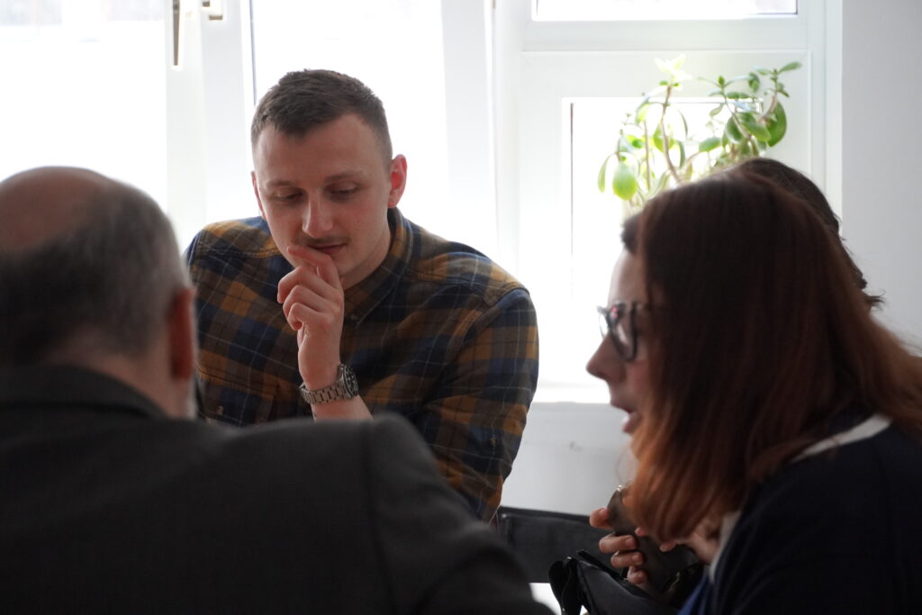 Mann in kariertem Hemd denkt in einer Besprechung nach, umgeben von mehreren Personen am Tisch.