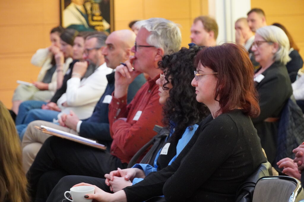 Zuhörende Erwachsene sitzen dicht beieinander bei einer Veranstaltung im Saal.