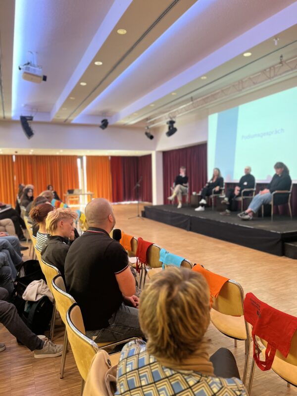 Paneldiskussion in einem Saal mit Publikum, Sprechern auf der Bühne und Projektion im Hintergrund.
