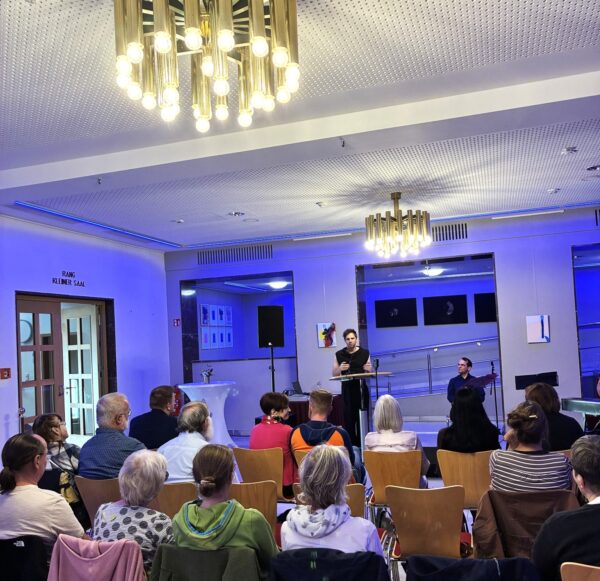 Vortrag vor sitzendem Publikum in einem kleinen Saal mit blauer Beleuchtung