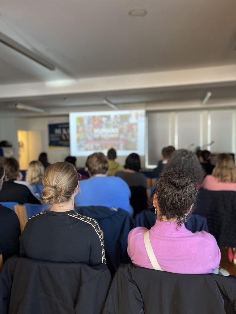 Publikum sitzt in einem Seminarraum und verfolgt eine Präsentation auf einer Leinwand