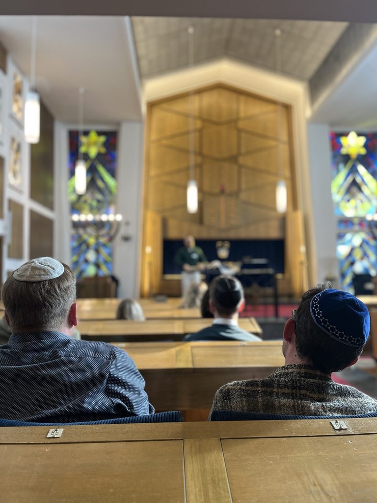 Männer mit Kippa sitzen in einer Synagoge und hören einem Vortrag vorn zu