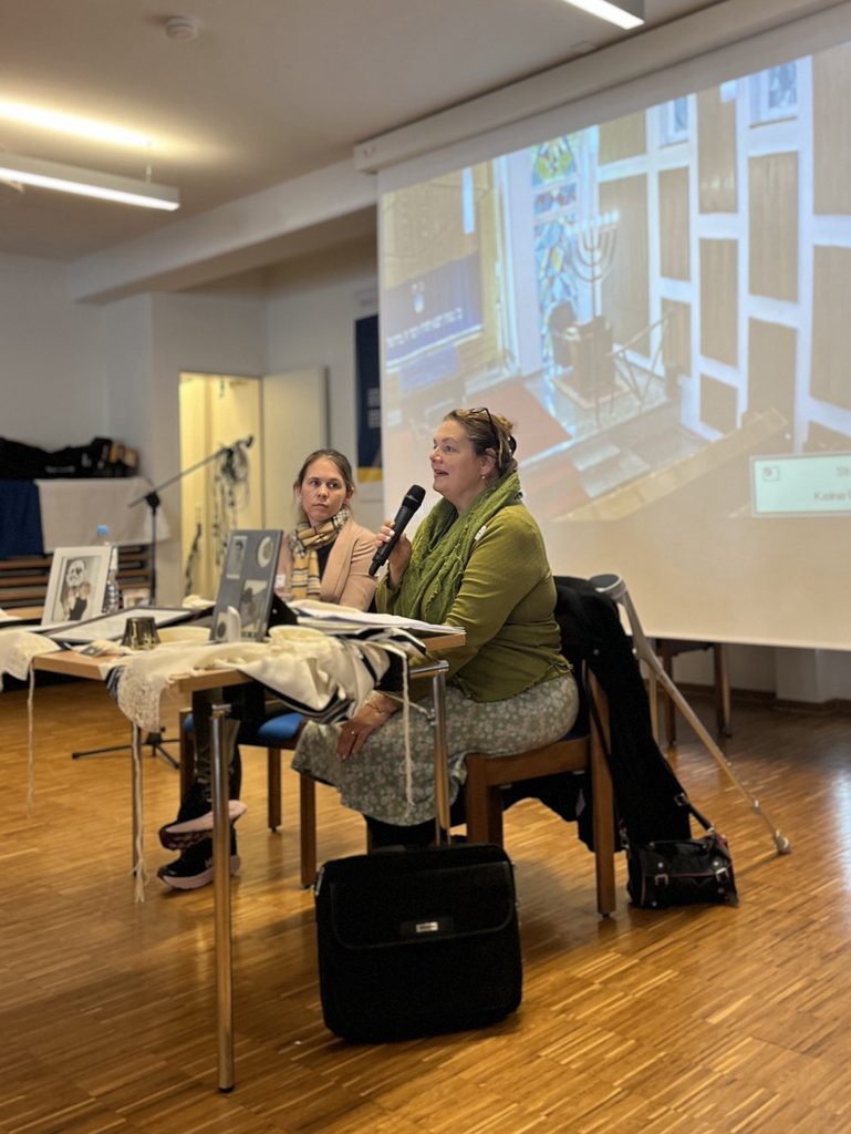 Zwei Frauen sprechen an einem Tisch bei einer Veranstaltung; eine hält ein Mikrofon, im Hintergrund läuft eine Projektion